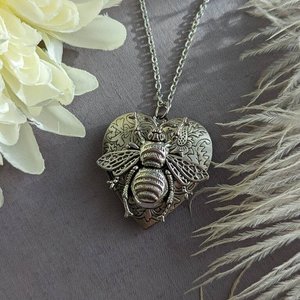 Silver Heart locket with floral design and large bee embellishment on top. Locket is displayed on a grey book with pale yellow flowers.