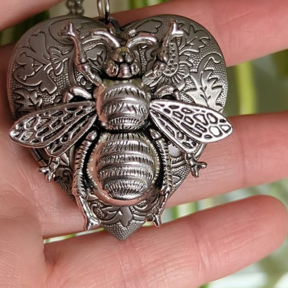 Silver Heart locket with floral design and large bee embellishment on top. Locket is being held in a hand.